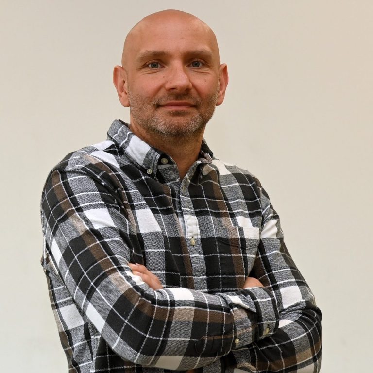 Mann mit kurzem Haar, stehend, in kariertem Hemd, mit verschränkten Armen.